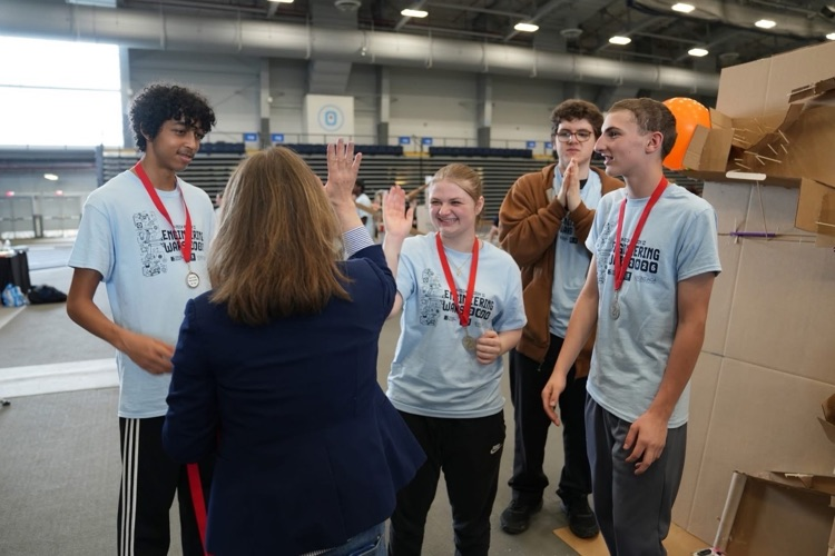 Students wearing medals high-five an adult in a large indoor arena during the Engineering Wars competition.