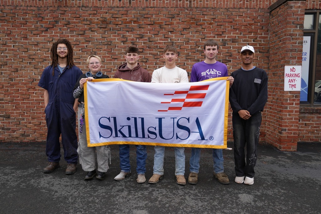 TST CTE juniors hold SkillsUSA flag outside of a brick building and smile at camera for group picture.