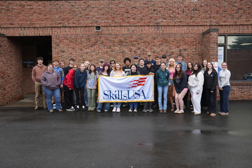 TST CTE seniors hold SkillsUSA flag outside of a brick building and smile at camera for group picture.