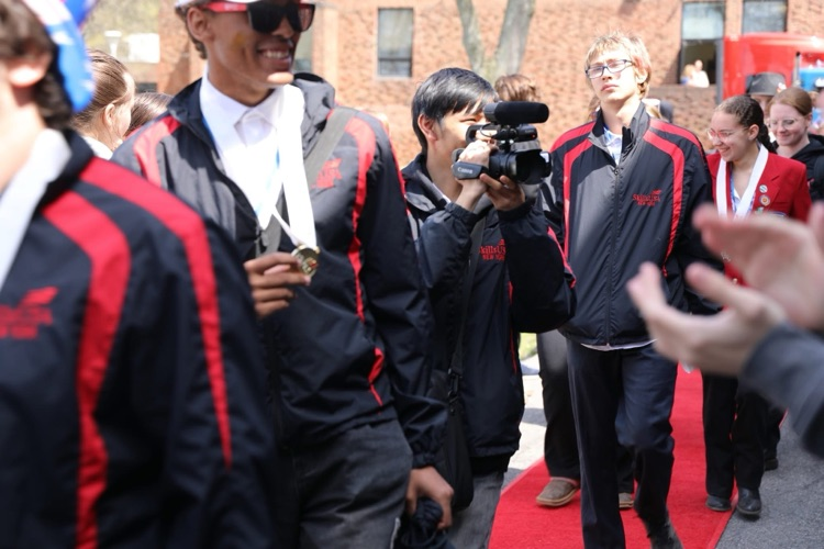 Group of SkillsUSA students walk together on the red carpet for the welcome back to TST parade, smiling, as one student films with a camera during the event.