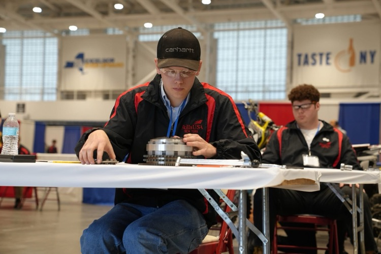 Student wearing safety glasses and a SkillsUSA jacket carefully assembles mechanical parts at a competition workstation, with another student working in the background.