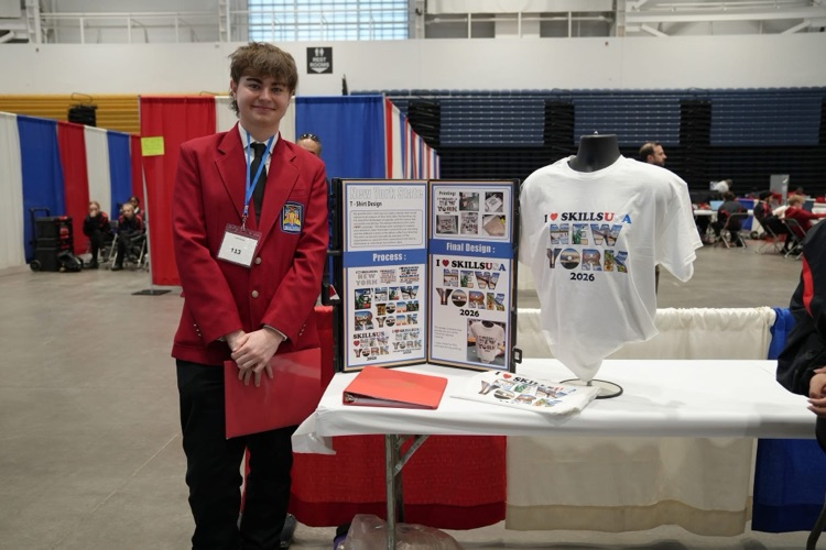Student in a red SkillsUSA blazer stands next to a display table featuring a project board and a T-shirt design presentation.
