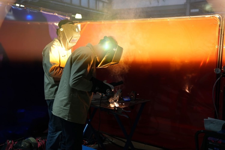 Two students wearing welding helmets work at a station, creating sparks and smoke as they weld metal behind a protective screen.