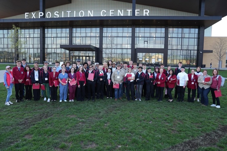 A large group of SkillsUSA students and advisors pose together outside the Exposition Center, holding folders and wearing red blazers and team apparel.