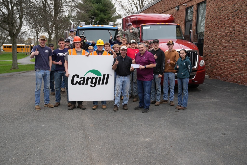 A large group of students and two adults pose in front of heavy trucks, smiling and giving thumbs up while holding a Cargill sign and a check.