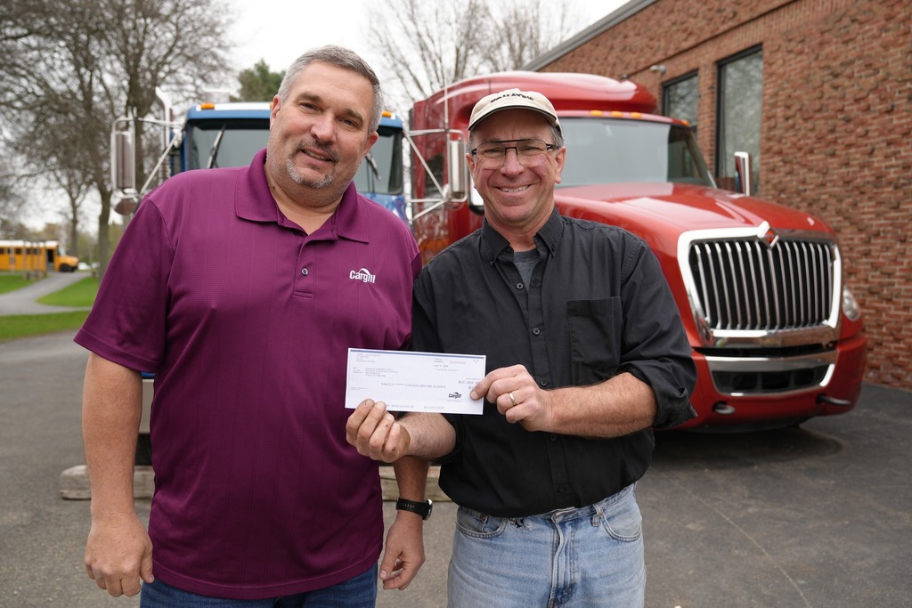 Two men stand in front of large trucks, smiling and holding a check together. One wears a Cargill polo shirt, and the other wears a black shirt and cap.
