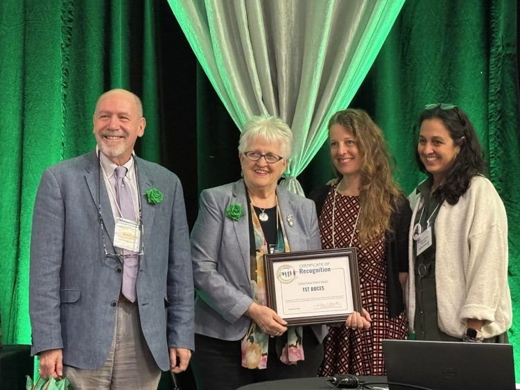  Dr. Dhundale, Ms. Jessica Hoff receive award on stage and smile with two other adults in front of a green curtain