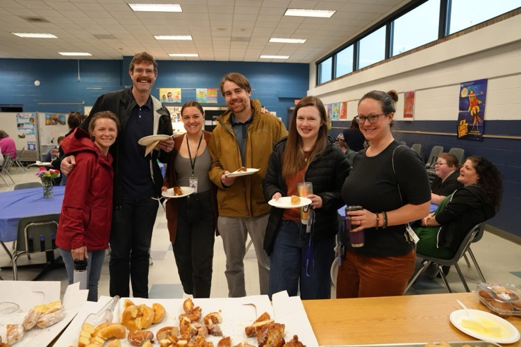 RAS teachers and staff stand together smiling with full plates in the cafeteria at the staff appreaciation breakfast