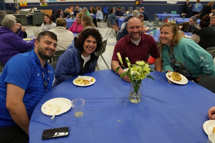 CTE teachers and staff stand together smiling  at a decorated blue round table and pretty flowers in the cafeteria at the staff appreaciation breakfast