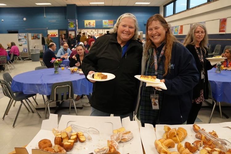 two staff members stand together smiling with full plates in the cafeteria at the staff appreaciation breakfast