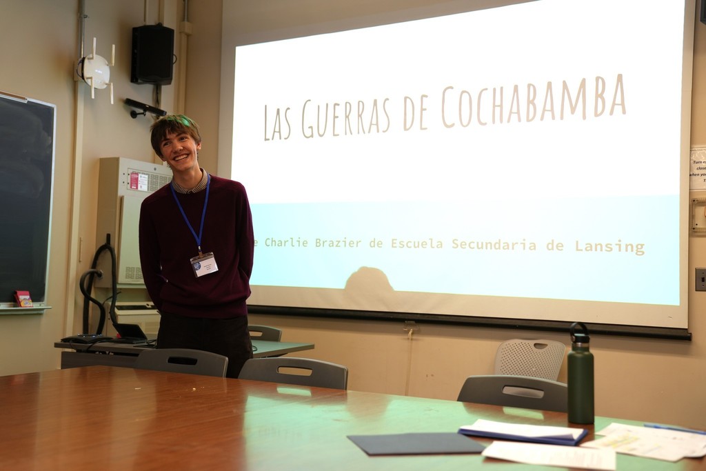 A student presents in Spanish during the Seal of Biliteracy event, standing in front of a presentation slide titled “Las Guerras de Cochabamba.”