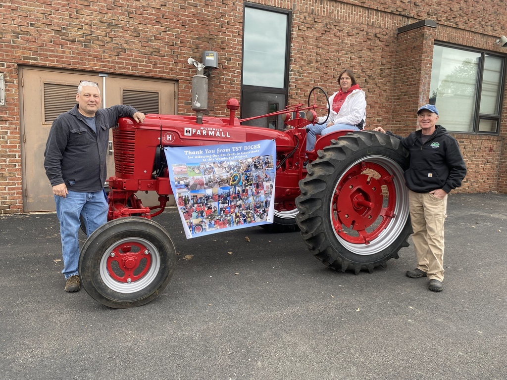The completed 1949 Farmall tractor is displayed outside TST BOCES with Jeff Brockway and its owners, Bill and Skip Wolfley, daughter Sarah. and a project banner showcasing photos from the student-led restoration process.