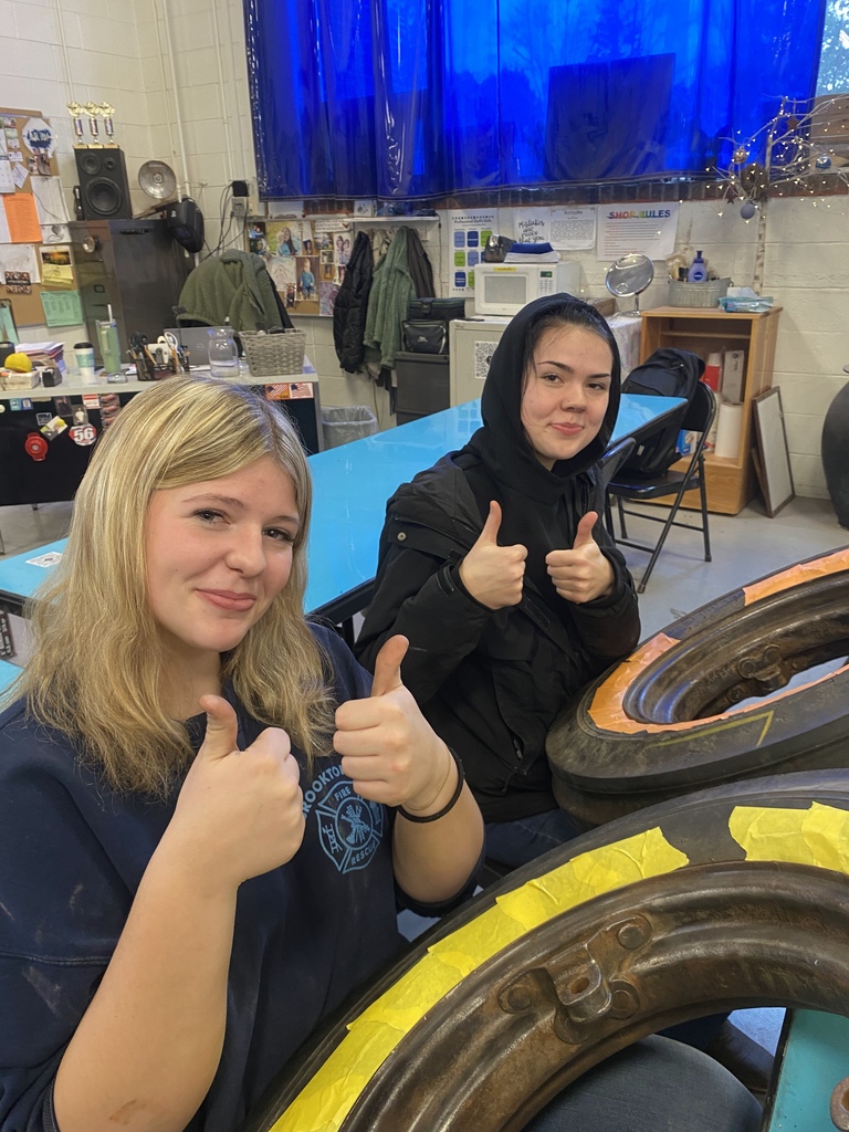 Two TST BOCES students give thumbs up while working on tractor wheel restoration in an Auto TBody classroom. Tractor rims and masking tape are visible in the foreground.