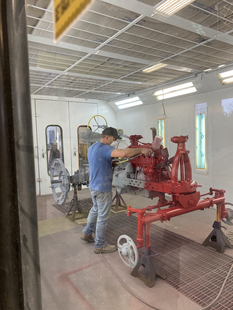 A student sprays bright red paint onto the frame and engine components of the 1949 Farmall tractor inside the Auto Body paint booth during the restoration process.