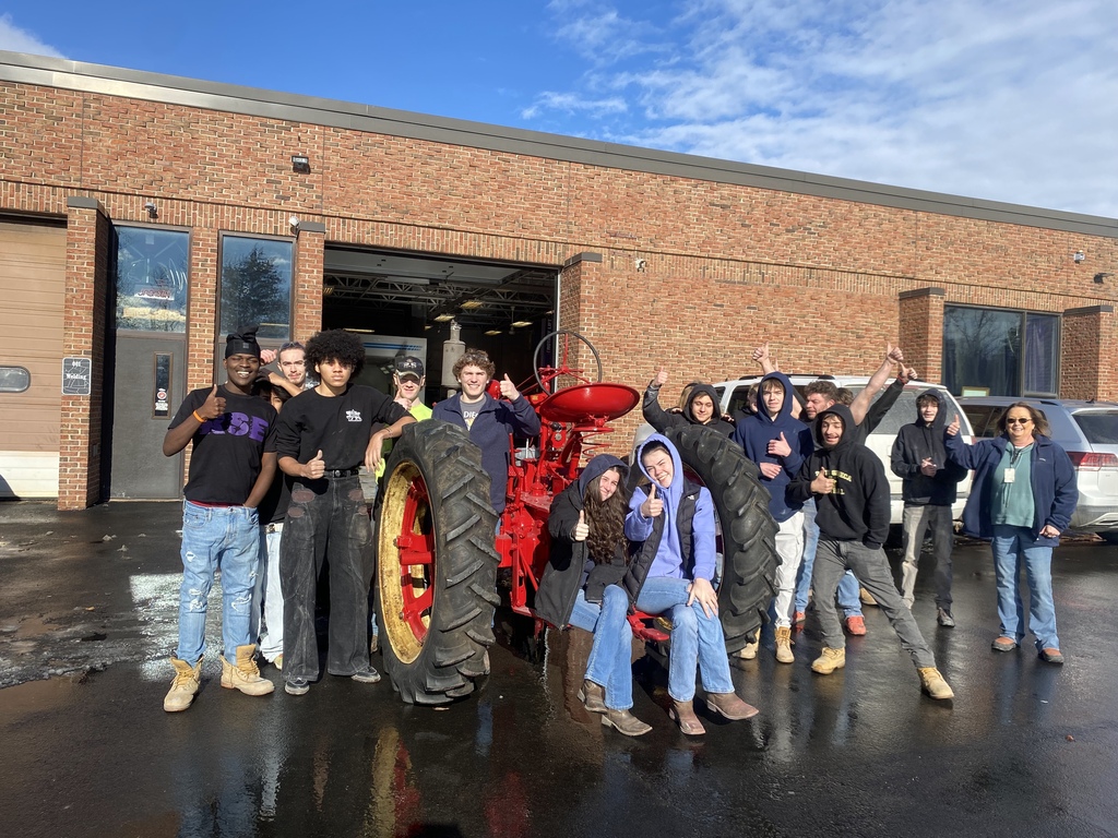 A group of TST BOCES Auto Body students and staff pose outside the CTE building with the partially restored bright red Farmall tractor, giving thumbs up and celebrating the project’s progress.