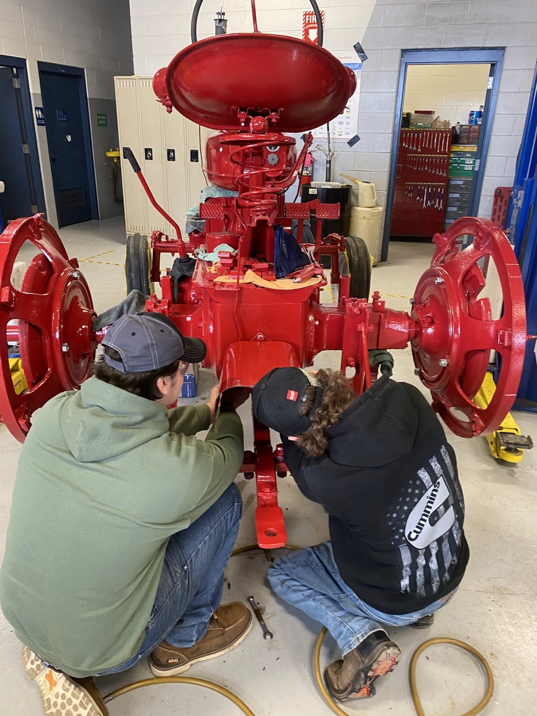Two students work underneath the rear section of the freshly painted red Farmall tractor while reassembling mechanical components in the Auto Tech shop.