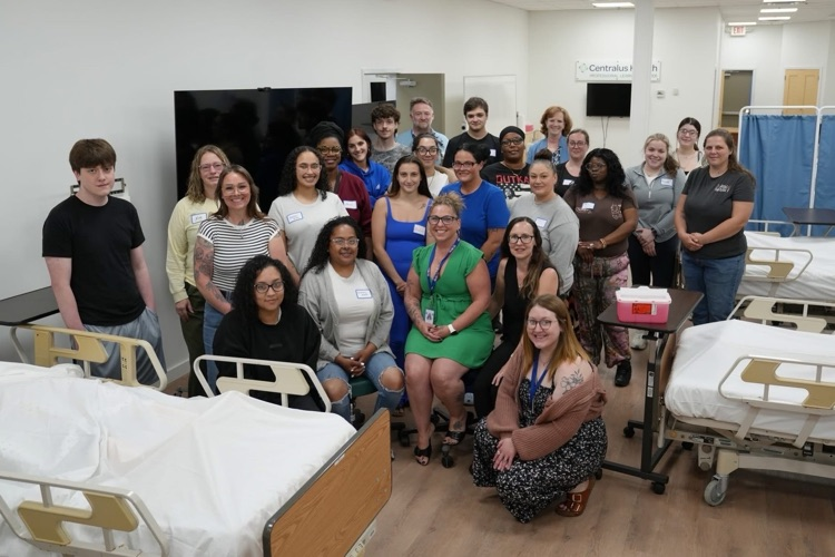 Group photo of the first TST BOCES Adult Education Practical Nursing Program cohort standing in a clinical training lab with hospital beds and medical equipment. Students and staff pose together on the first day of the program.