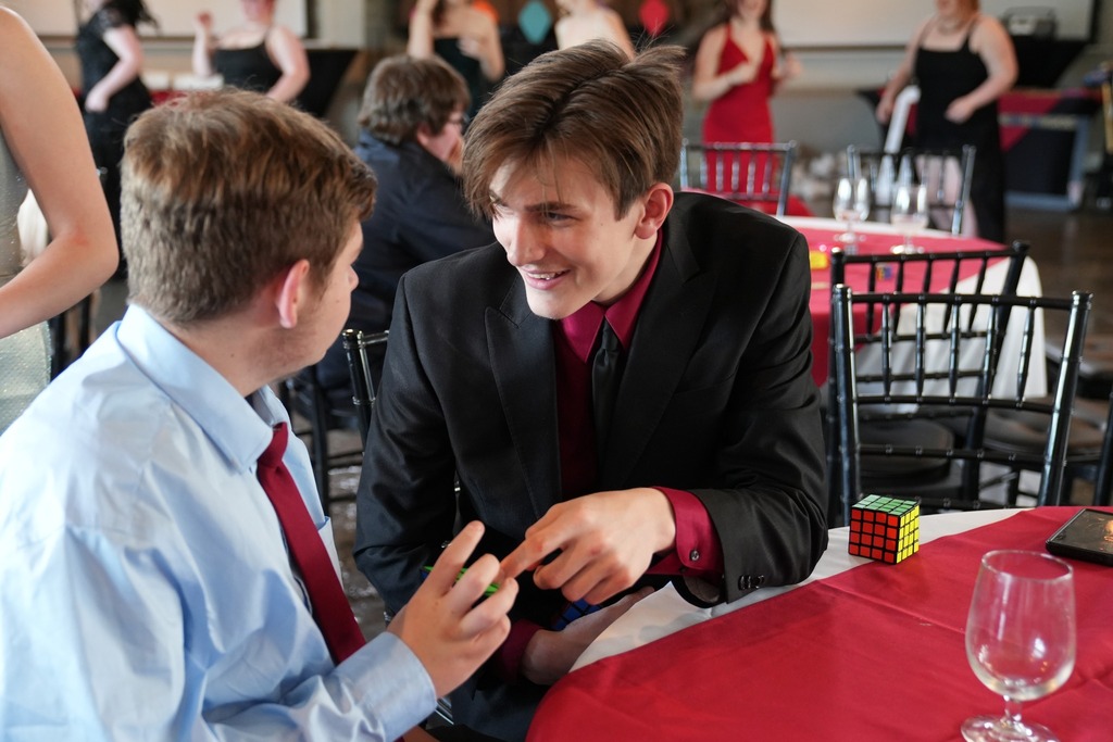Two students in dress shirts and ties sit at a table talking and solving Rubik’s cubes during the dinner dance.