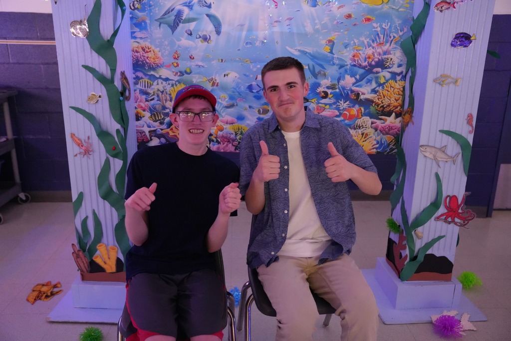 Two students sit in front of an underwater-themed photo backdrop and give thumbs up while posing for a picture.