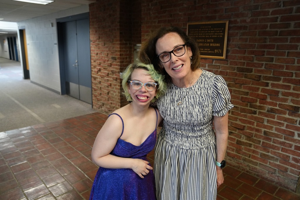 Student in a sparkly blue dress smiles beside an adult in a striped dress in a hallway at prom.