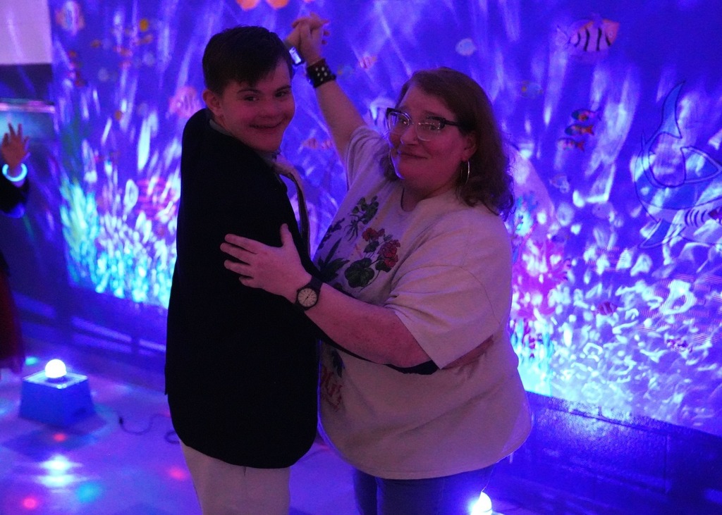 Student and staff member dance together in front of an underwater-themed backdrop with glowing blue and purple lights during prom.