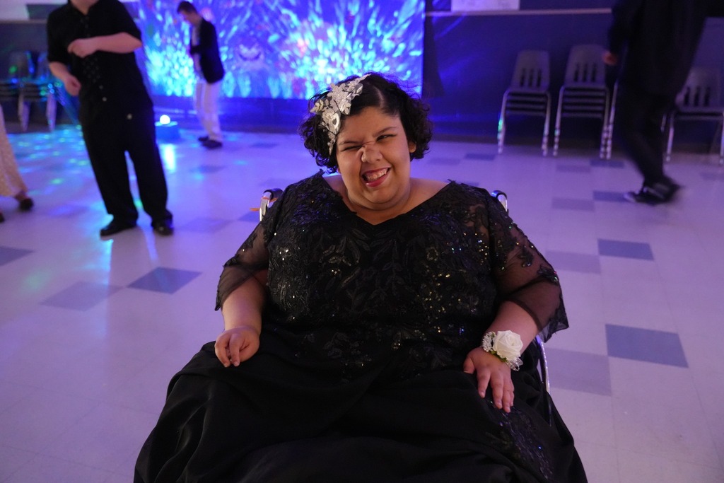 Student in a black formal dress smiles and poses in a wheelchair on the dance floor during the Under the Sea themed Exceptional Education Prom, with colorful blue lights glowing in the background.