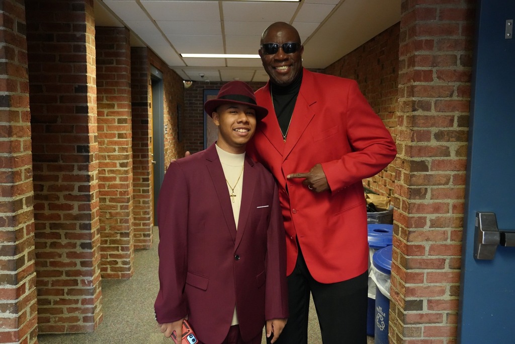 A student smiles with a staff member dressed in coordinated red outfits pose together in a brick hallway during the Exceptional Education Prom.