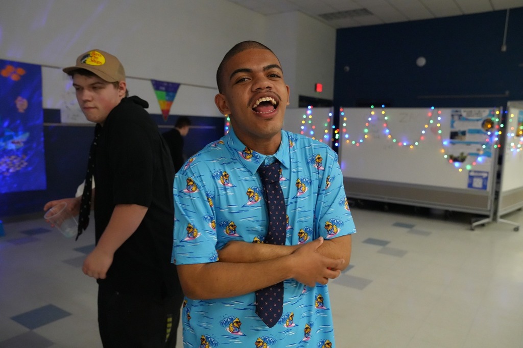 Student wearing a blue tropical-print shirt and tie smiles on the dance floor during prom while another student stands nearby.