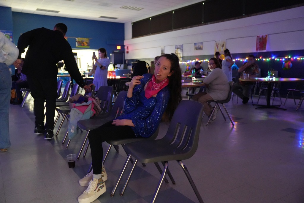 Student in a blue sequined top and pink scarf sits and smiles in the decorated prom venue while other attendees gather in the background.