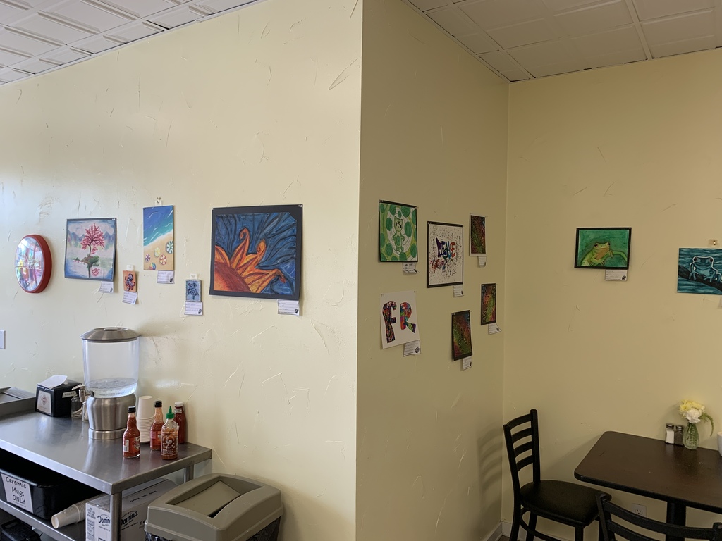 Corner view of a café showcasing artwork by TST BOCES RAS students. Colorful paintings and mixed-media pieces are displayed on adjoining walls above tables, chairs, and a condiment station.