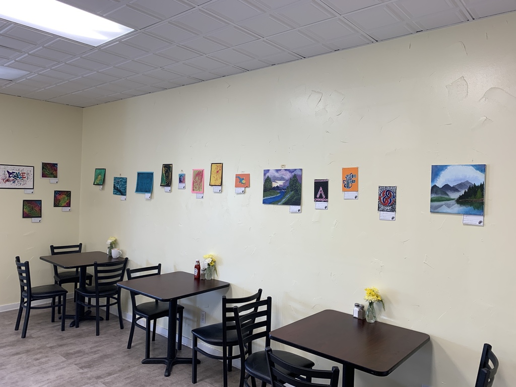 A collection of student artwork hangs on a café wall, featuring landscapes, abstract pieces, graphic designs, and nature-inspired paintings. Several café tables are arranged below the display.