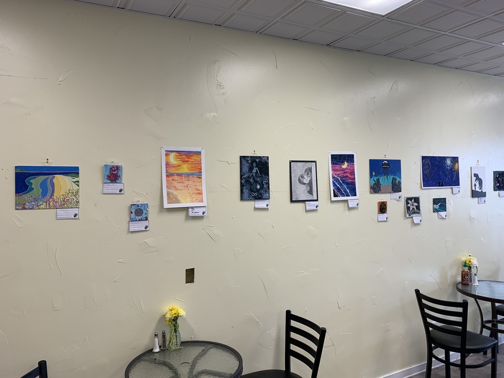 Wide view of a café wall displaying a student art exhibit featuring colorful paintings, drawings, and mixed-media pieces created by TST BOCES RAS students. Small café tables with flowers sit beneath the artwork.
