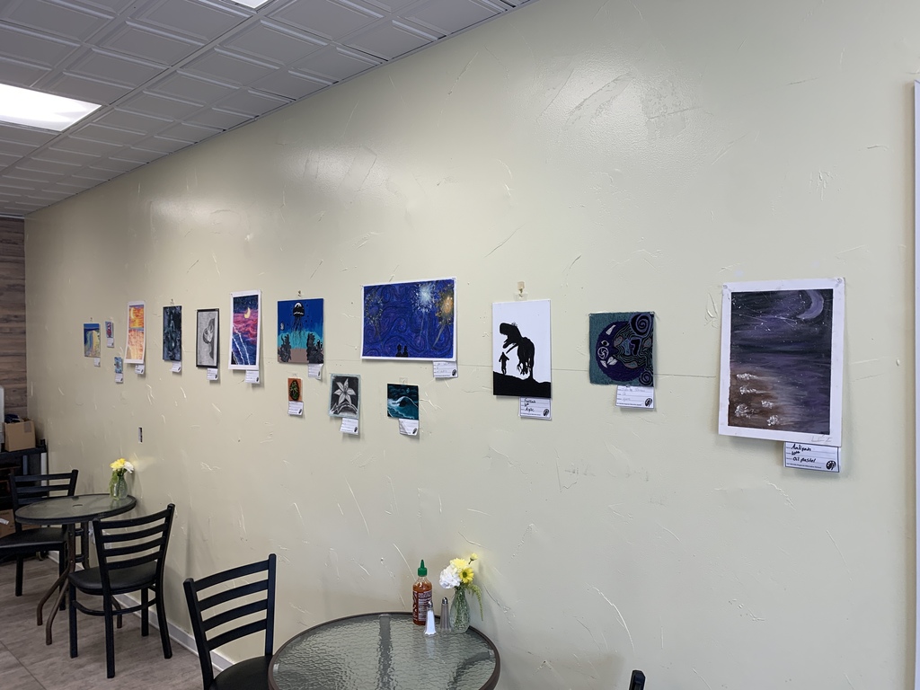 Artwork by TST BOCES RAS students displayed along a café wall, including paintings of night skies, ocean scenes, abstract designs, and silhouettes. Café tables and chairs are visible in the foreground.