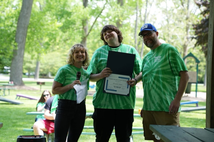 World of Work from Food Service student stands between two staff members while holding a certificate during an outdoor awards presentation at the annual WOW Picnic.