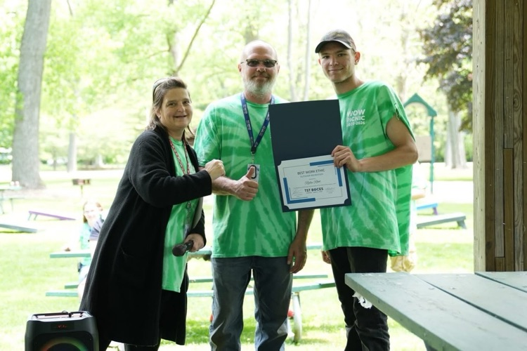 World of Work student stands between two staff members while holding a certificate during an outdoor awards presentation at the annual WOW Picnic from Outdoor Power Class