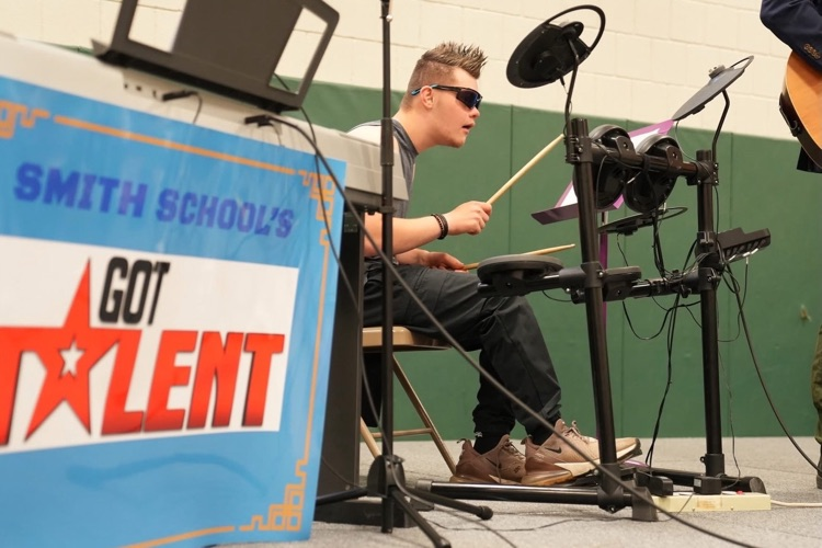 Student wearing sunglasses plays an electronic drum set during Smith School’s Got Talent while seated on stage beside a sign displaying the event logo.