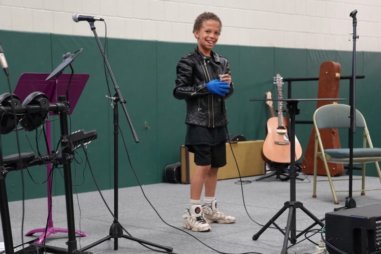 Student in a black jacket and gloves stands smiling at a microphone on stage, preparing to perform during Smith School’s Got Talent.