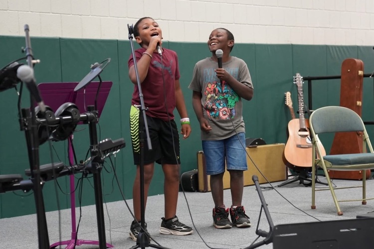 Two students sing together into handheld microphones on stage, smiling as they perform a duet during the talent show.