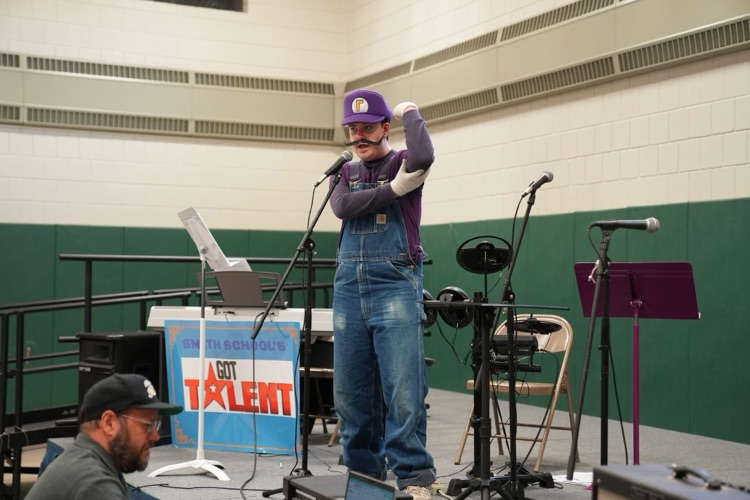  Person dressed as the video game character Waluigi performs on stage during Smith School’s Got Talent, speaking into a microphone while striking a dramatic pose. Musical equipment, microphones, and a Smith School’s Got Talent sign are visible on stage in the school gymnasium.