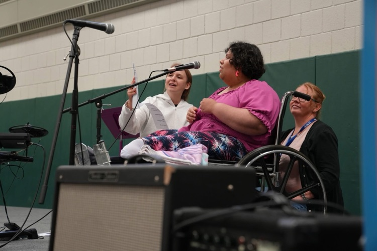Student seated in a wheelchair sings into a microphone while another staff member holds lyric sheets nearby. A staff member sits beside them on stage. 