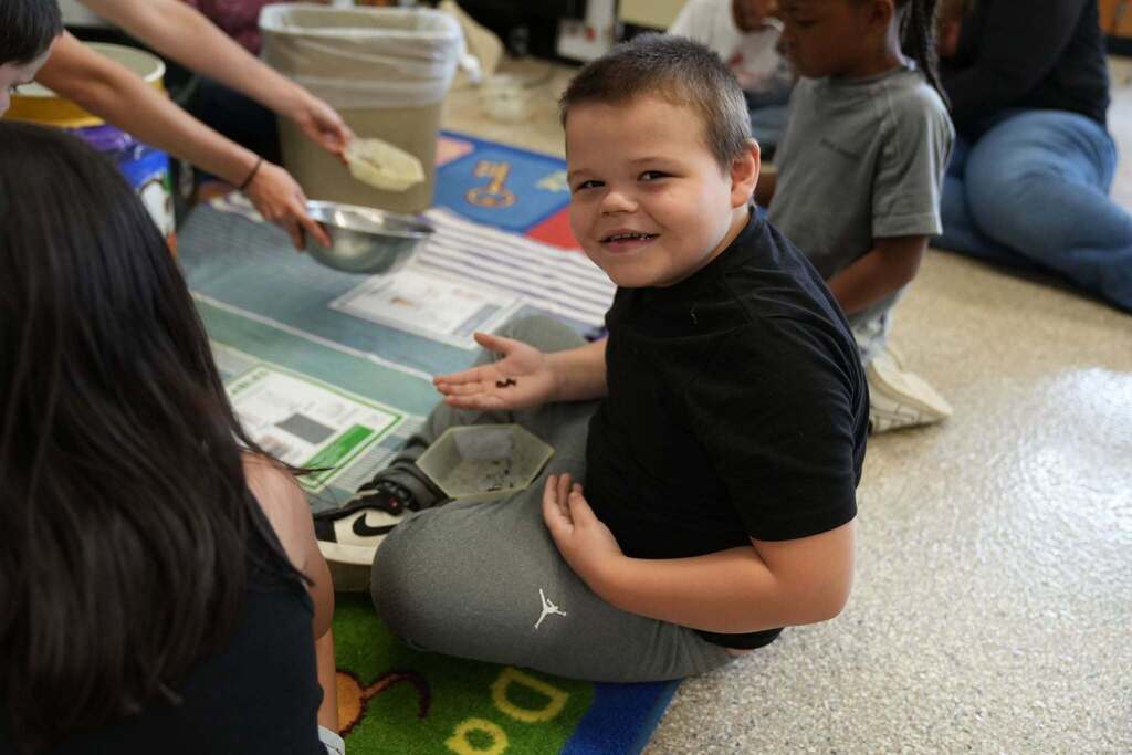 A smiling student holds a small worm in their hand while taking part in a hands-on composting activity with classmates.
