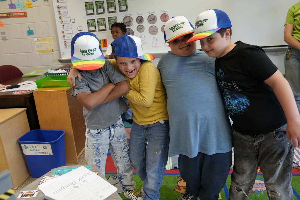 Four students pose together wearing colorful "Compost Is Cool" hats during a classroom celebration of their composting and environmental learning.