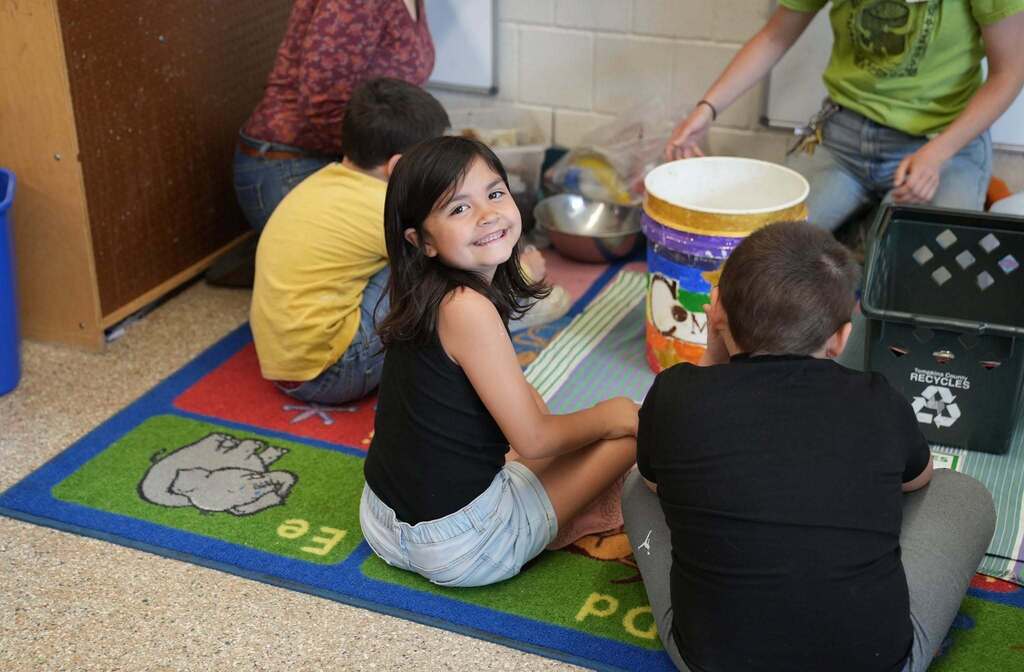 A student smiles toward the camera while participating in a hands-on composting activity with classmates and educators.