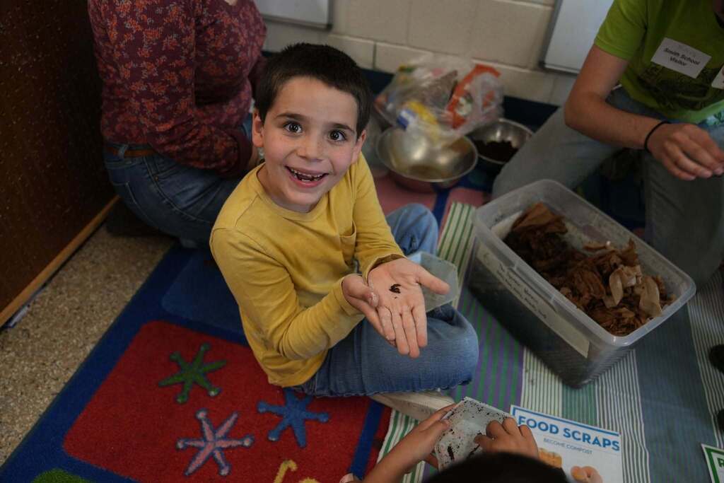 A student smiles while carefully holding a worm during a composting lesson, with compost bins and food scrap materials nearby.