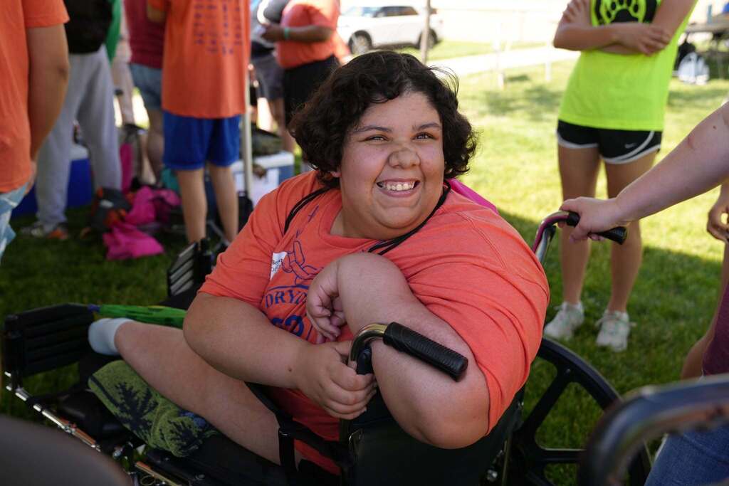 Smiling student seated in a wheelchair enjoys a break between events at the Dryden Games while surrounded by classmates and volunt