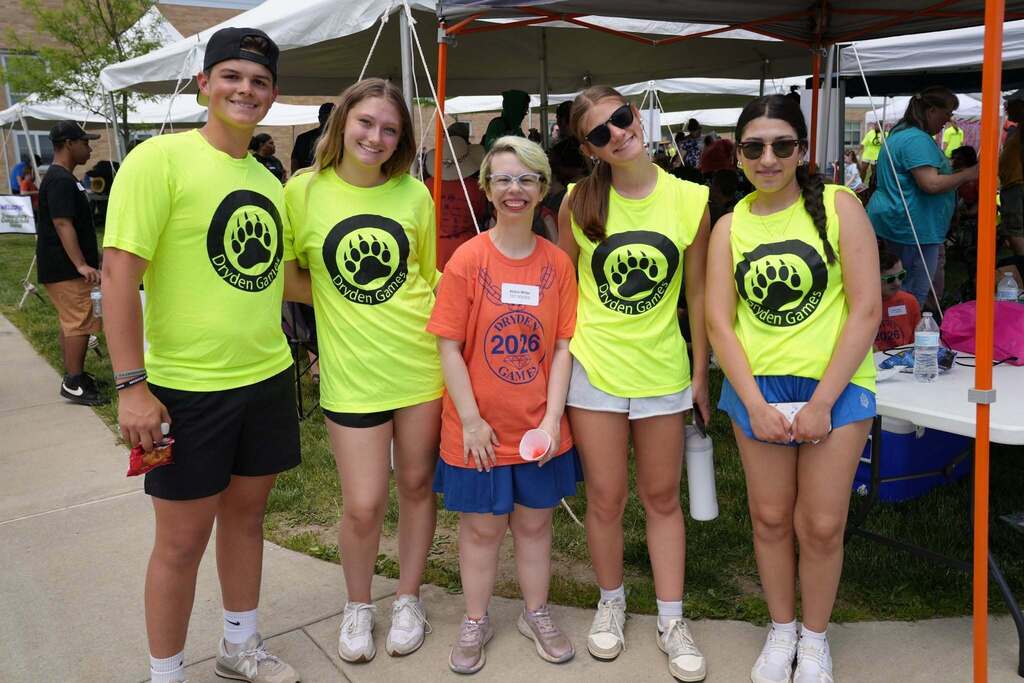 Student participant stands smiling with four student volunteers wearing Dryden Games shirts under an event tent.