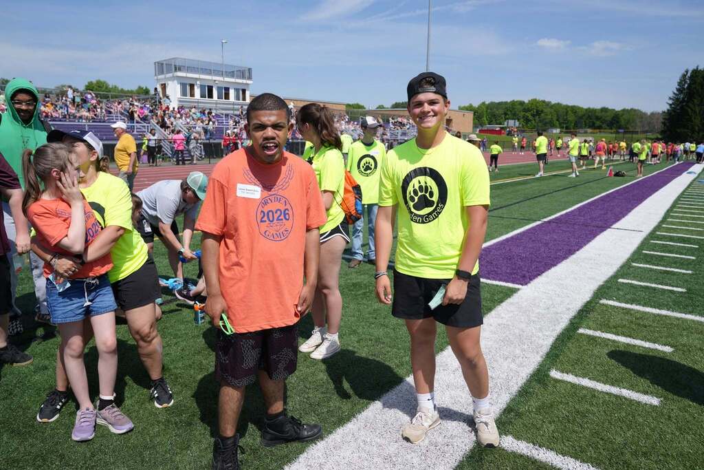 Student participant and volunteer pose together on the football field during the Dryden Games with athletes and spectators in the background.