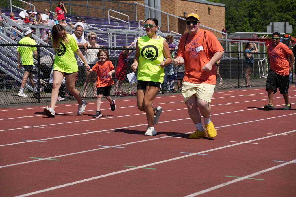 Student participants and volunteers jog together on the track during a relay event at the Dryden Games while spectators cheer from the bleachers.