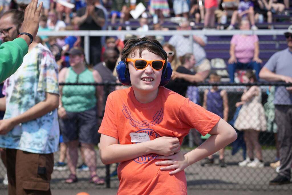 Student wearing orange sunglasses and hearing protection stands on the field during a Dryden Games event with spectators in the background.