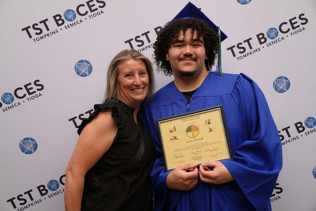 Graduate in a blue cap and gown proudly displays a Circle of Courage Award certificate while posing with a family member.